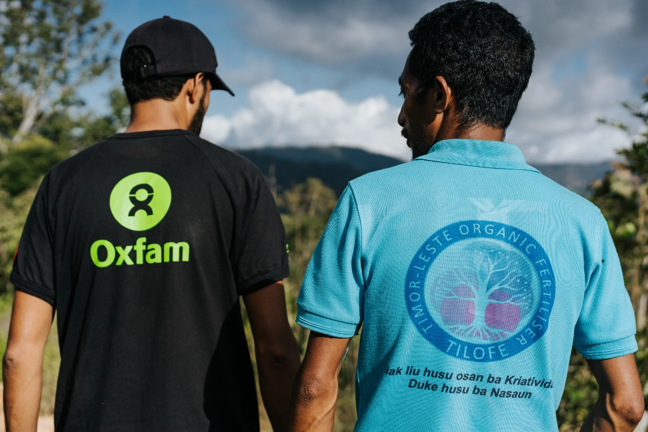 Two people, one wearing a black shirt with a green Oxfam logo and one in a blue shirt with a TILOFE logo, walk on a farm and appear to be in conversation with their backs facing us.