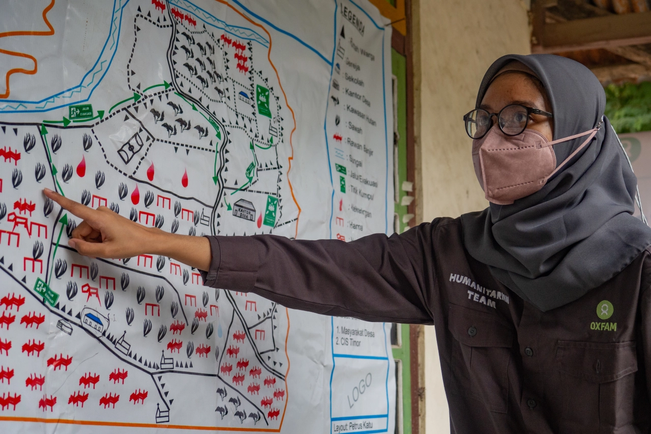 A woman in a hijab, mask, and glasses points at a portion of a map.