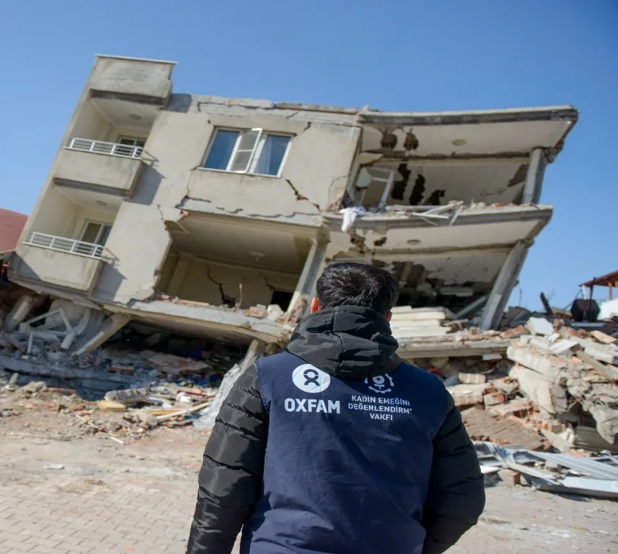 A man dressed in a hooded puffer jacket and a blue vest with an Oxfam logo and KEDV logo stands in front of a collapsed house with his back to the camera.
