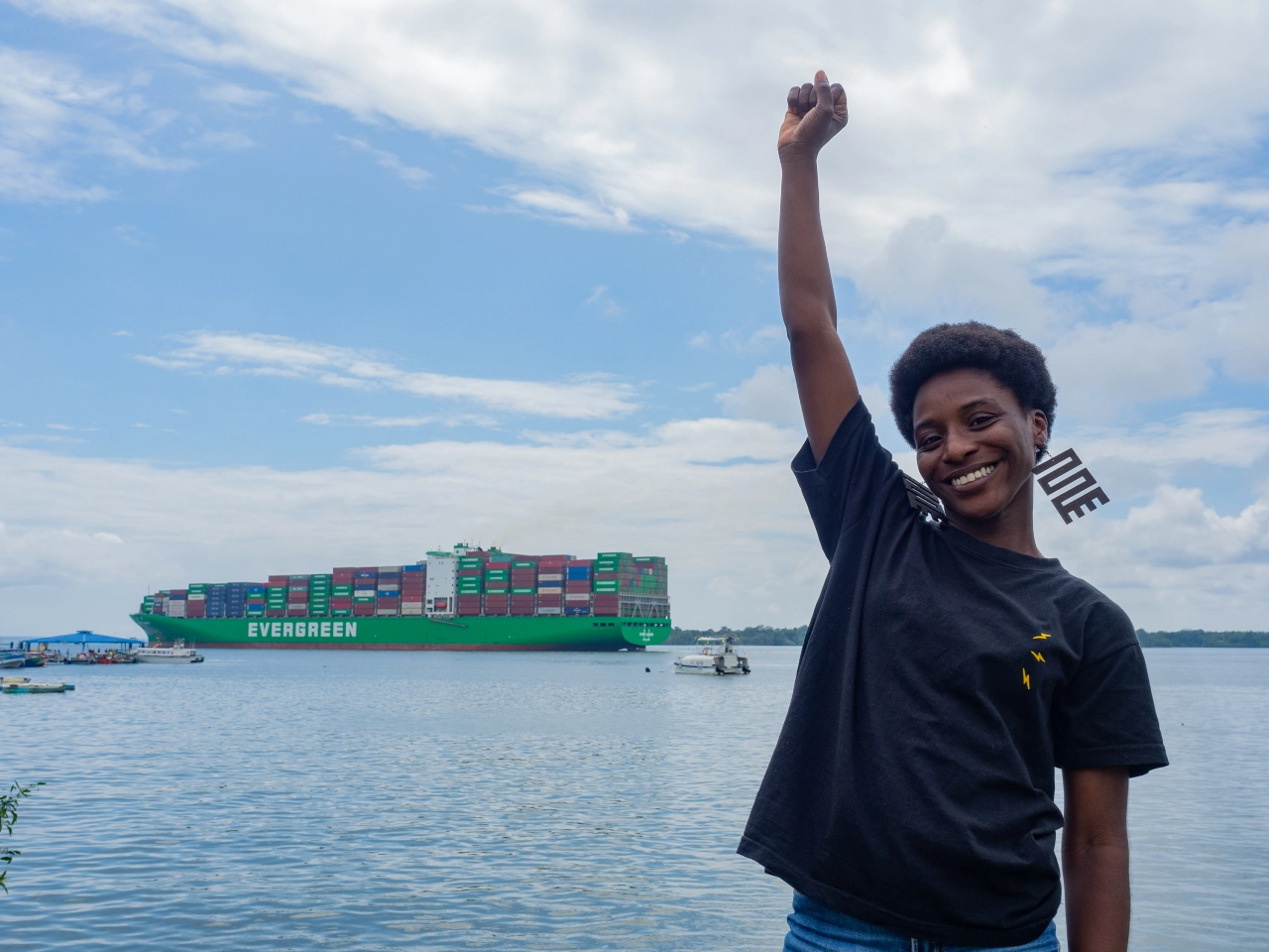 An activist stands in front of a large body of water with her right fist raised triumphantly. To her left, in the background, is a large green ship transporting containers.